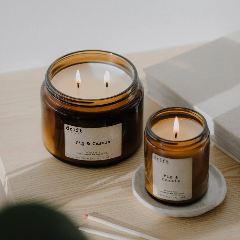 Two lit candles in amber jars with labels on a wooden surface.