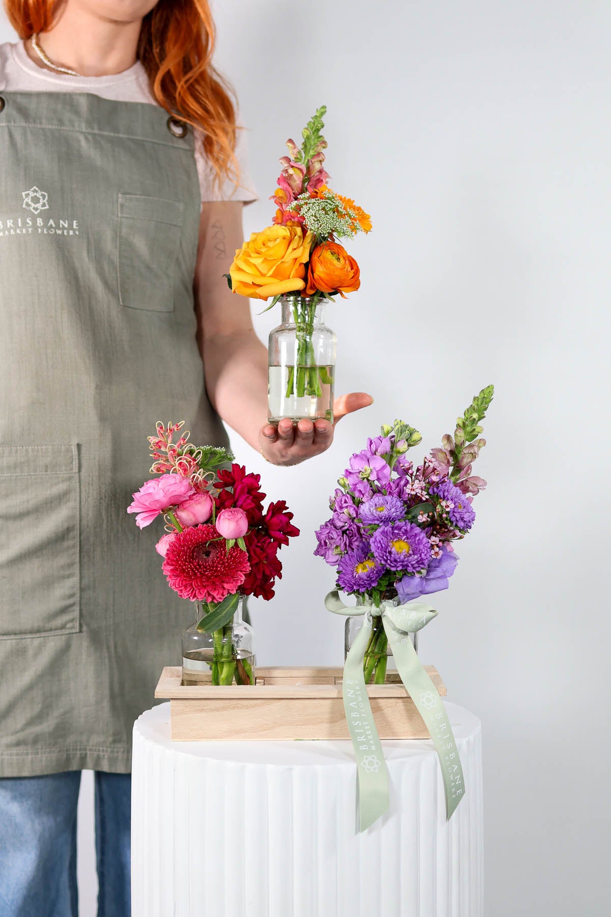 Person holding a small glass vase with flowers, surrounded by other floral arrangements on a white surface.