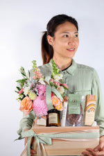 Woman holding a gift box hamper with choclates, candle, hand cream with flowers and other items against a white background