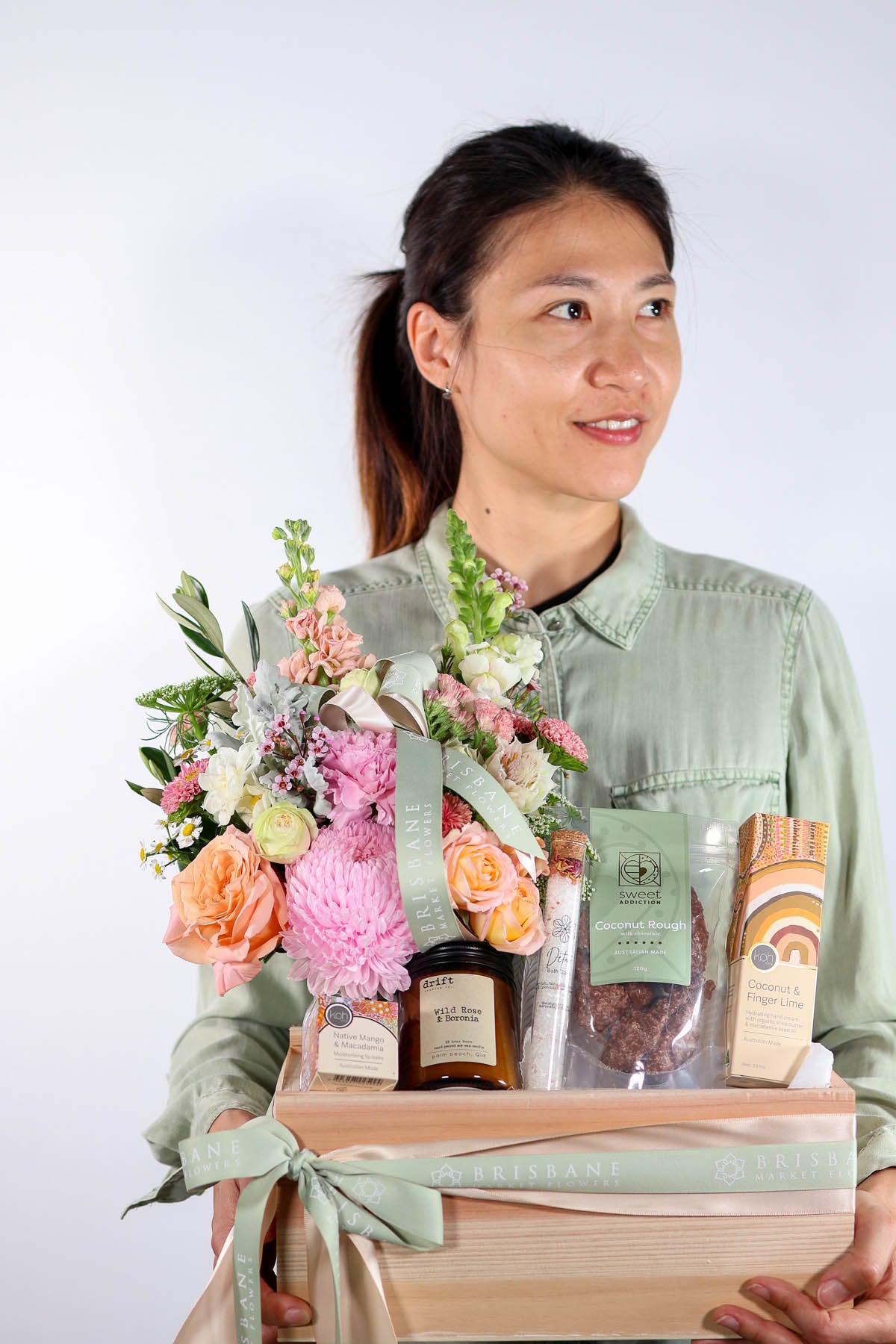 Woman holding a gift box hamper with choclates, candle, hand cream with flowers and other items against a white background