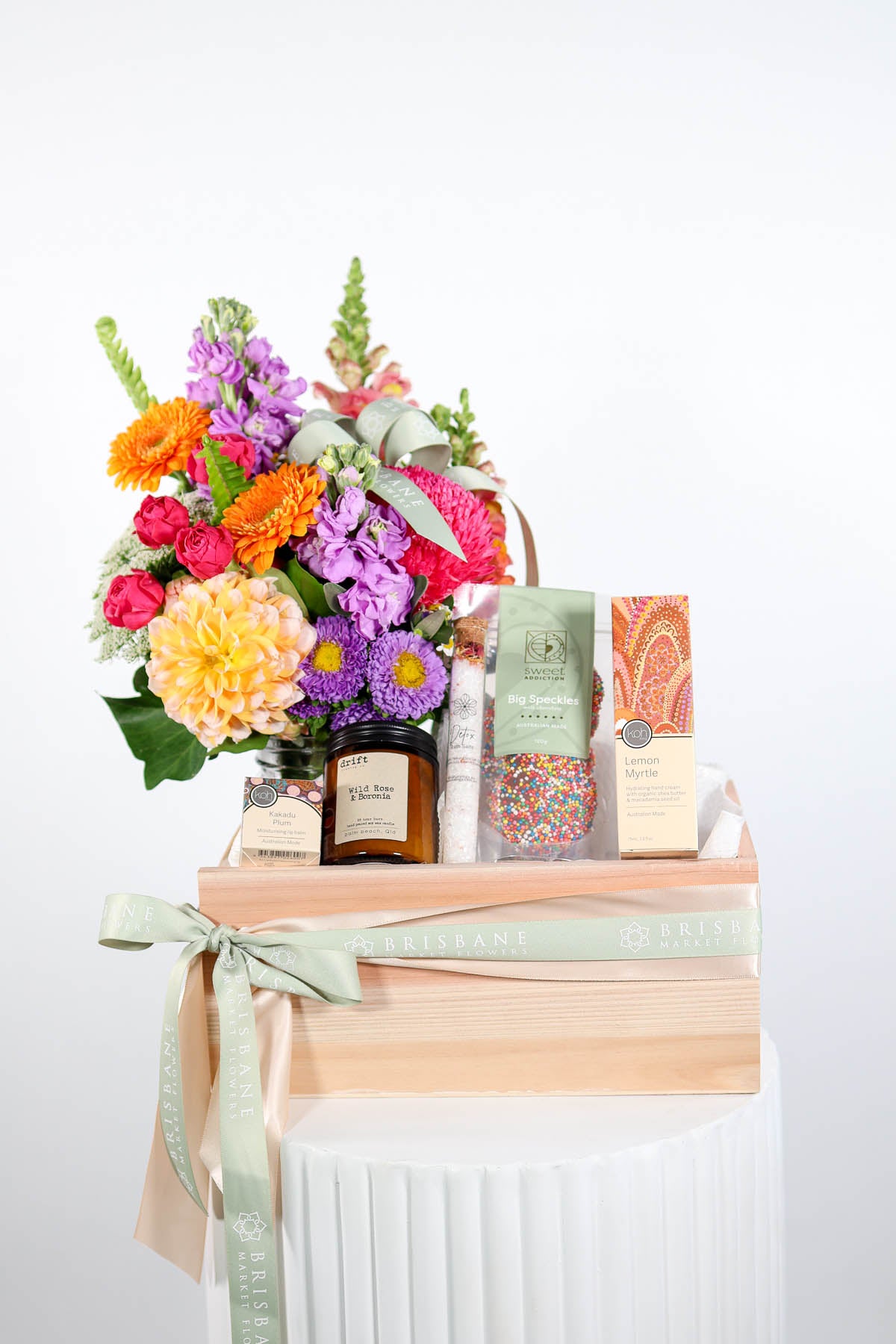 Bouquet of flowers with decorative gift items like a candle, hand cream, speckle chocolates and other items in a bamboo crate on a white stand against a white background