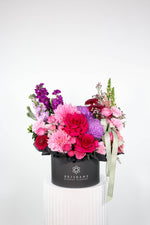 Colourful pink and purple flower arrangement in a black box with 'Brisbane Market Flowers branding, on a white background