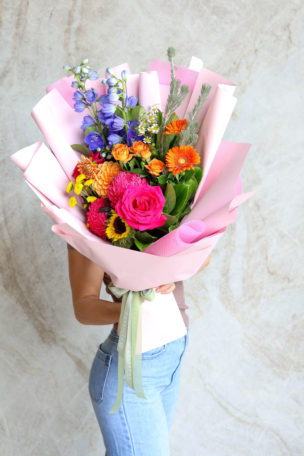 Person holding Graduation Bright Summer Bouquet featuring pink, orange, yellow & blue flowers to show size and front-facing style