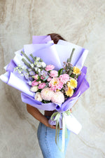 Pastel graduation bouquet with soft  blooms wrapped in lilac ruffle paper, front-facing presentation.