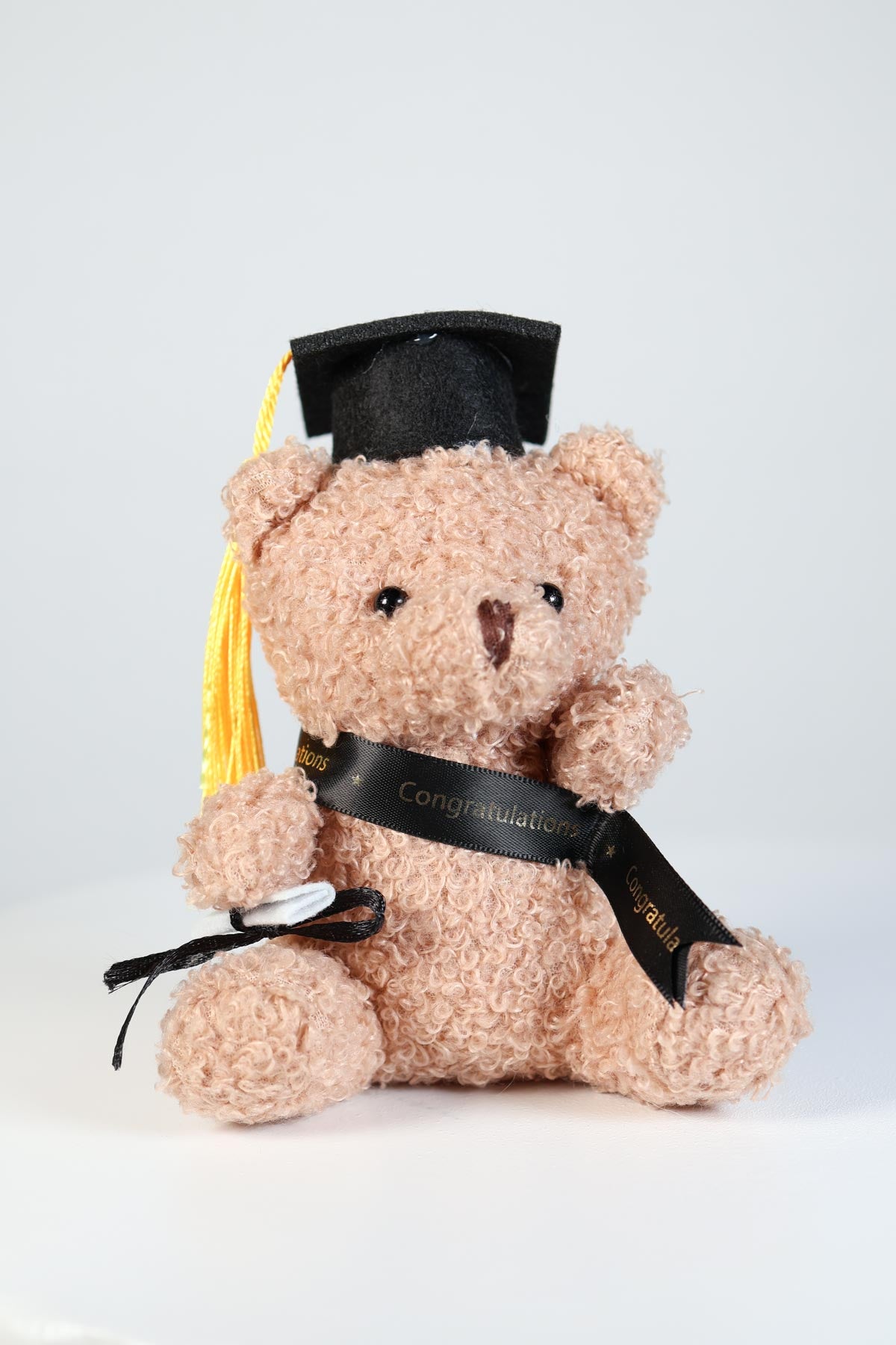 Graduation-themed teddy bear with a black cap and yellow tassel on white background.