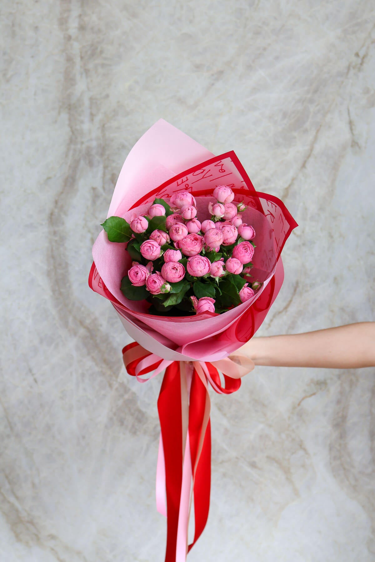 Bouquet of pink flowers wrapped in red paper with a ribbon against a marble background.