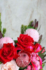 Bouquet of red roses, pink proteas, and other flowers with a pink ribbon on a light background.