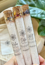 A hand holding three tubes of Naturalogy Botanical Bath Salt tubes with various botanicals and mineral salts visible