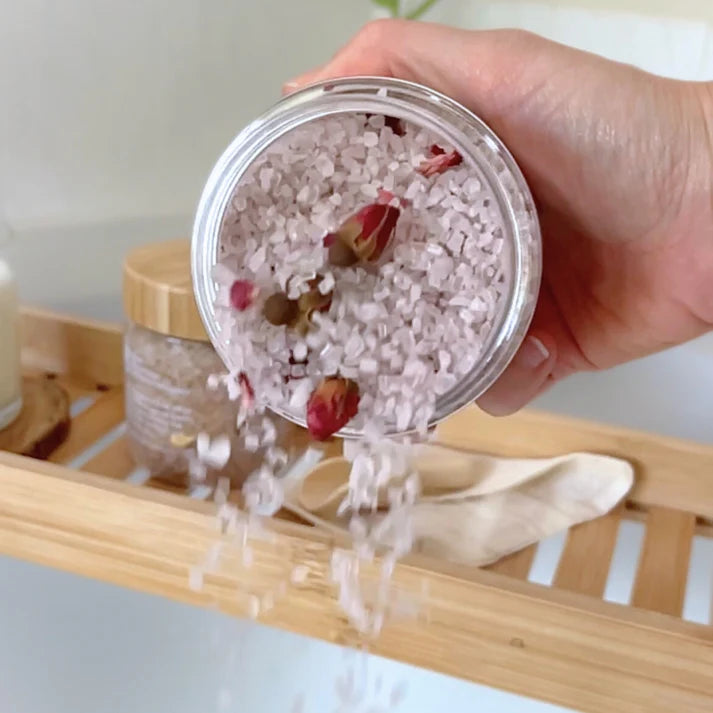 Hand holding a jar of bath salts with flowers, with a wooden shelf in the background.