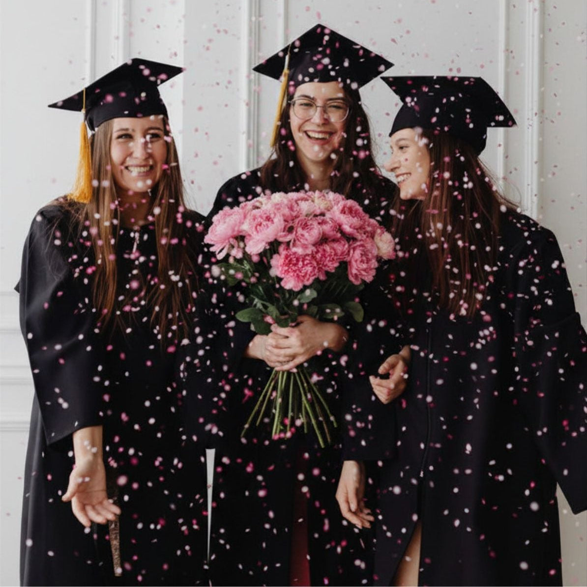 Three women in graduation gowns and caps with a bouquet of pink flowers.