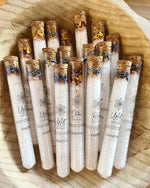 A collection of Naturalogy Botanical Bath Salt tubes with various botanicals and mineral salts visible, displayed in a wooden bowl.