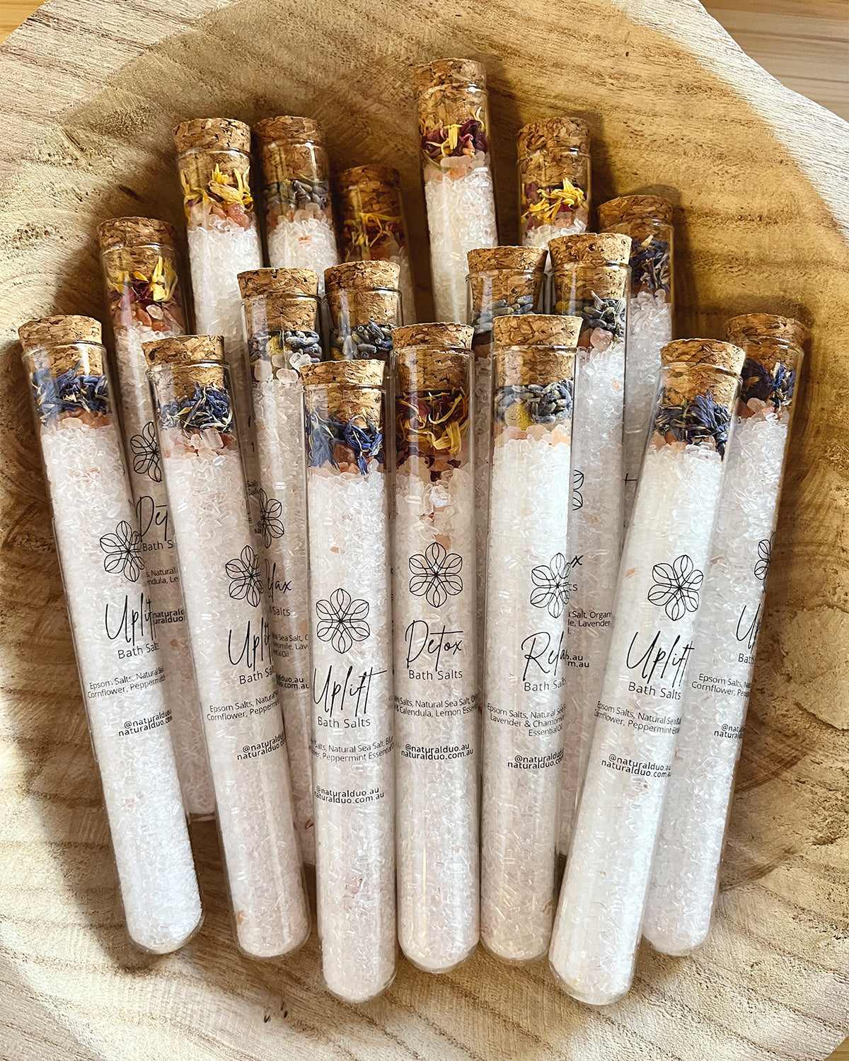 A collection of Naturalogy Botanical Bath Salt tubes with various botanicals and mineral salts visible, displayed in a wooden bowl.