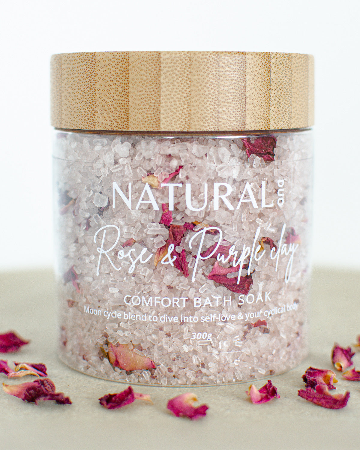 A jar of Naturalogy Rose & Purple Clay Comfort Bath Soak, with pink bath salts visible and scattered rose petals around it.