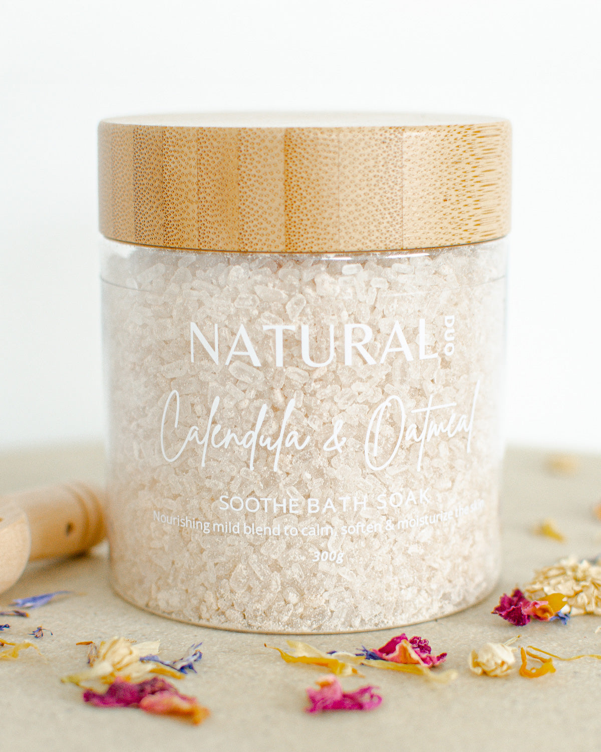 A jar of Naturalogy Rose & Purple Clay Comfort Bath Soak, with pink bath salts visible and scattered rose petals around it.
