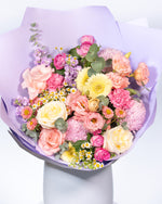 A floral bouquet featuring a mix of blush pink, creamy yellow, and soft purple flowers, including roses and chrysanthemums, wrapped in a purple paper.