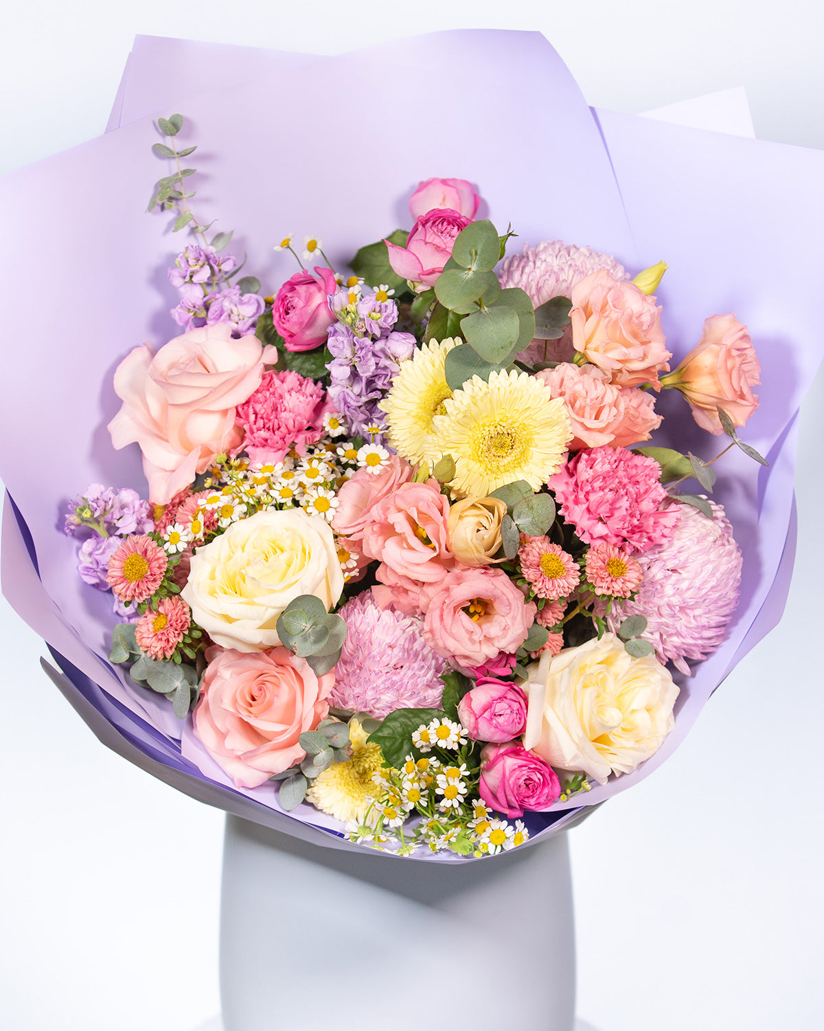A floral bouquet featuring a mix of blush pink, creamy yellow, and soft purple flowers, including roses and chrysanthemums, wrapped in a purple paper.
