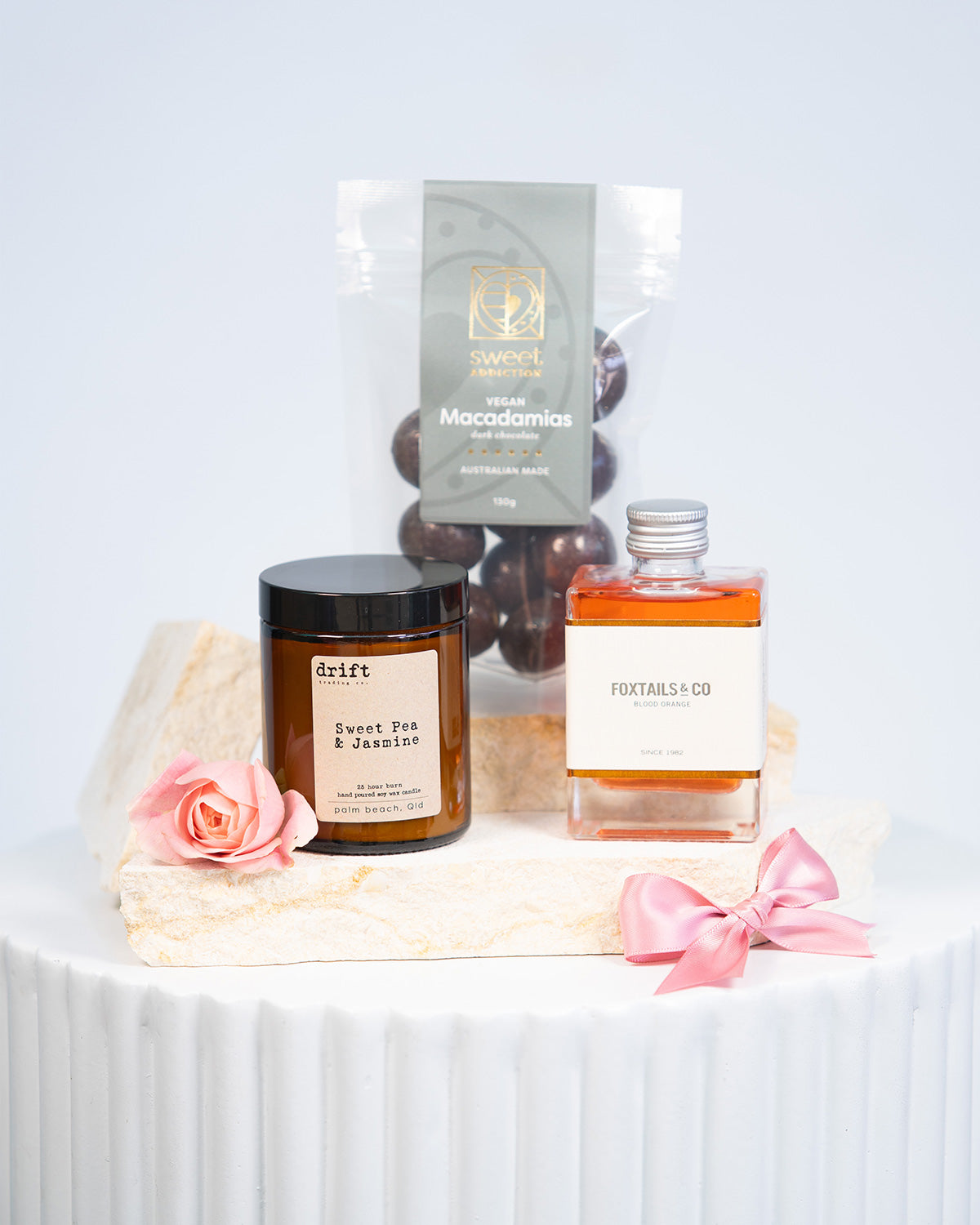 A gift hamper setup with an assortment of products including a candle, a packet of chocolates, and a small bottle of hand cream, all presented on a white surface with a pink bow.