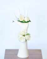 A wrist corsage featuring off-white spray roses with foliage, attached to a mannequin hand, holding a matching rose buttonhole.