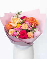 A seasonal bouquet featuring a bright mix of flowers including shades of pink, gold, apricot, and orange arranged in a round fashion, wrapped in pink paper.