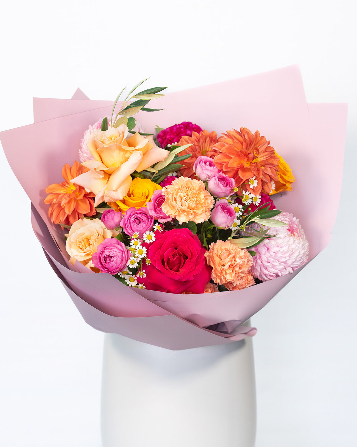 A seasonal bouquet featuring a bright mix of flowers including shades of pink, gold, apricot, and orange arranged in a round fashion, wrapped in pink paper.